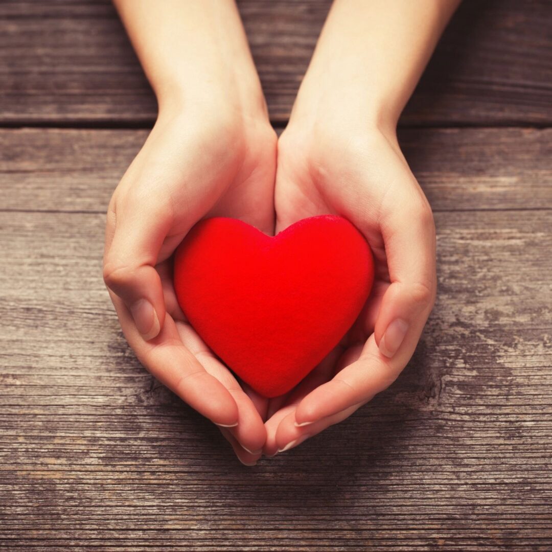 Hands cupping red heart on wooden background